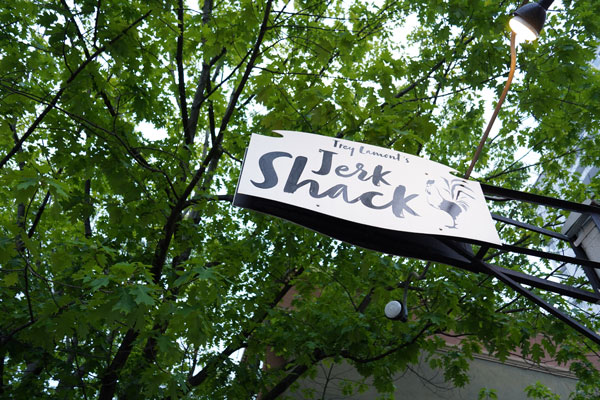 Sign for "Trey Leonard's Jerk Shack" against a backdrop of green tree leaves and a cloudy sky.
