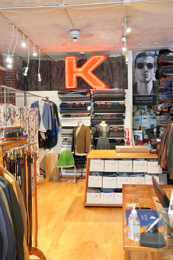 A clothing store interior with neatly stacked fabric rolls, assorted jackets on racks, and a counter with drawers. A red neon "K" sign is mounted on the wall. A mannequin wears a jacket and shirt. A large poster with sunglasses is on one wall.