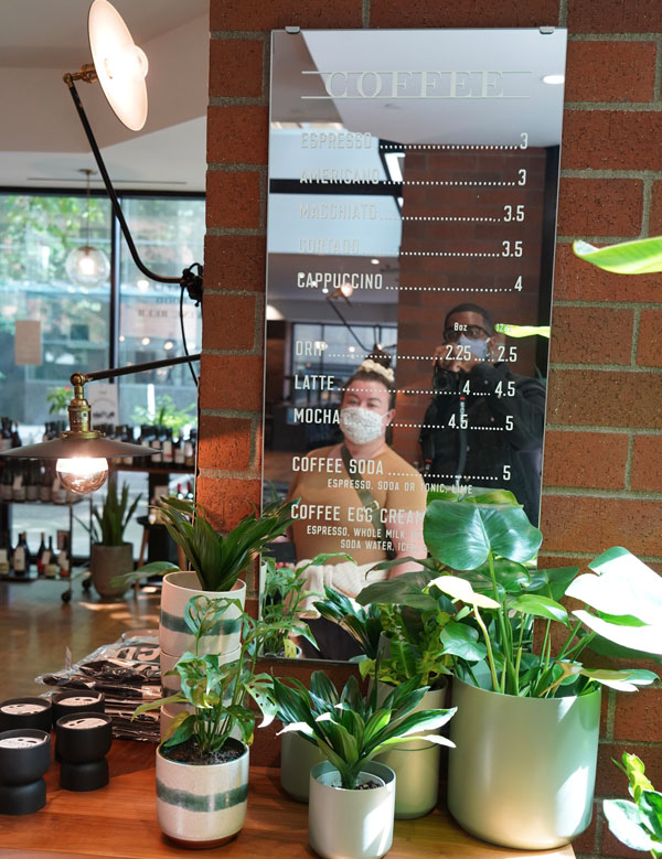 A cozy coffee shop interior featuring a menu board with coffee prices on a brick wall. Potted plants and decorative lights are in the foreground. A customer in a yellow jacket and mask is reflected in the mirror.