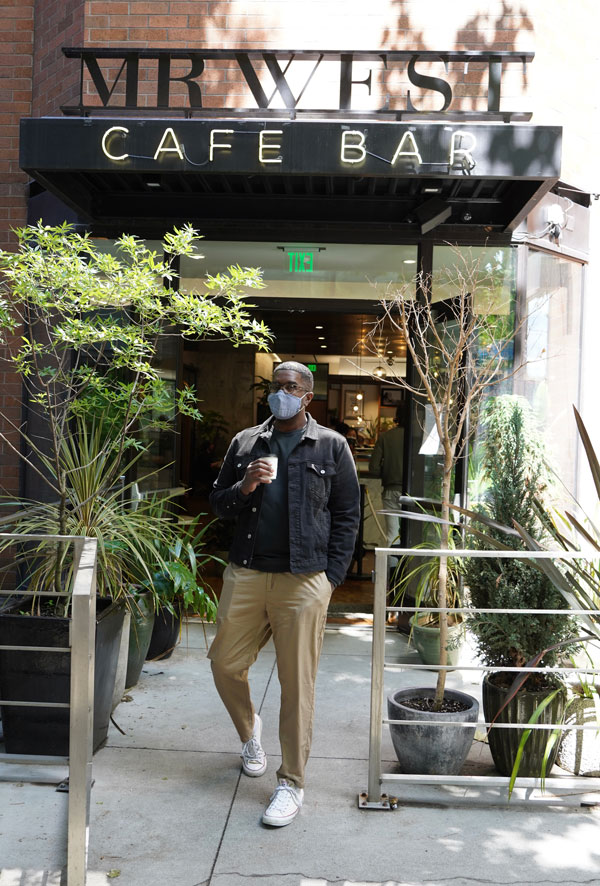 A person wearing a mask and sunglasses stands holding a cup outside Mr. West Cafe Bar. They wear a dark jacket, beige pants, and white sneakers. The cafe’s sign is visible above, and the entrance is surrounded by plants.