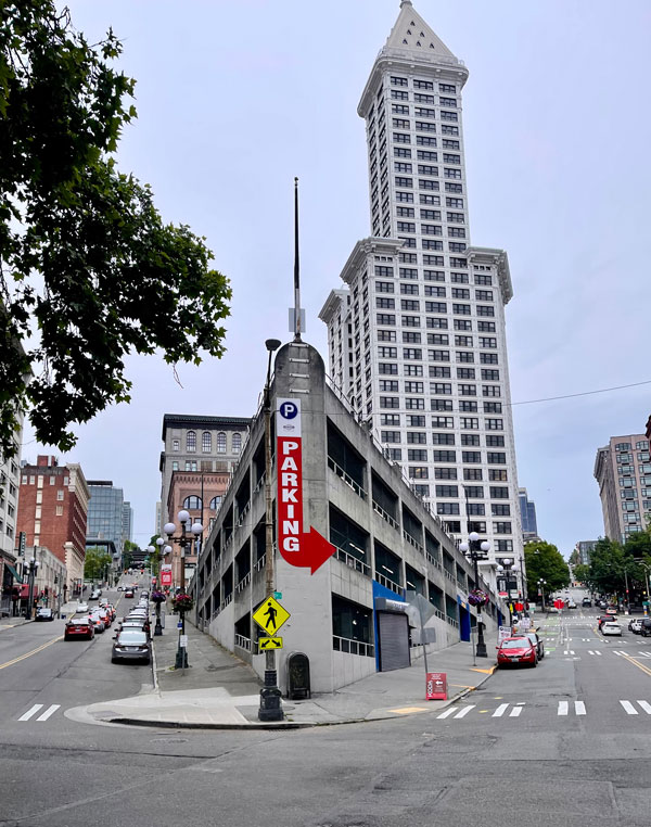 A multi-story parking garage with a red "Parking" sign sits at a street intersection, with a tall skyscraper in the background. Cars are on the road, and a pedestrian crossing sign is visible. Trees line the street, and the sky is overcast.