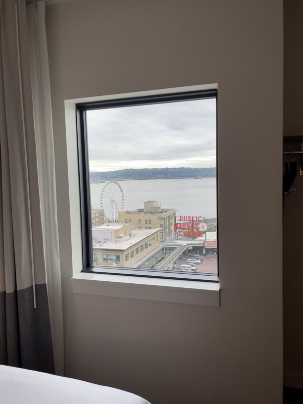 View from a window showing a cloudy sky over a waterfront. A Ferris wheel and a red neon sign reading "PUBLIC" are visible on top of a building. The scene includes buildings and a body of water in the background.