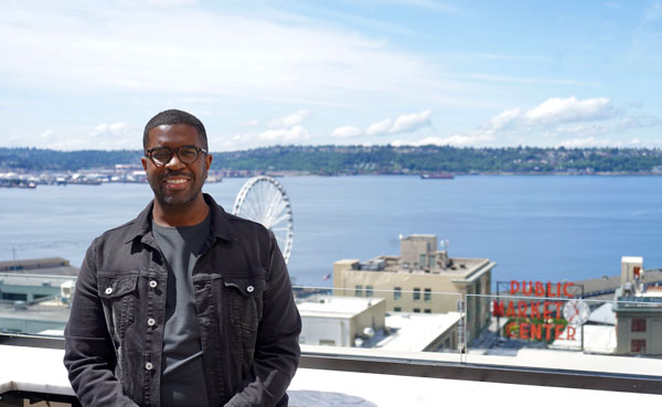 A person smiling, standing in front of a scenic view of a waterfront city. Behind them, there's a Ferris wheel and buildings, including a sign for "Public Market Center." The sky is clear with a few clouds.