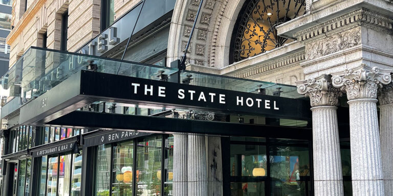 Entrance of The State Hotel, featuring a grand archway with intricate stonework and columns. A black awning displays the hotel name in bold white letters, and there's a glimpse of the street and cityscape in the background.