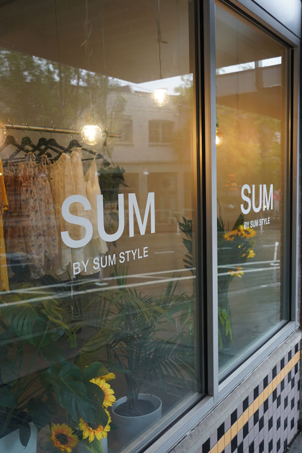 Storefront window with the words "SUM BY SUM STYLE" visible. Inside, clothing is displayed on racks, and sunflowers are placed at the base. The reflection of trees and buildings is seen on the glass.