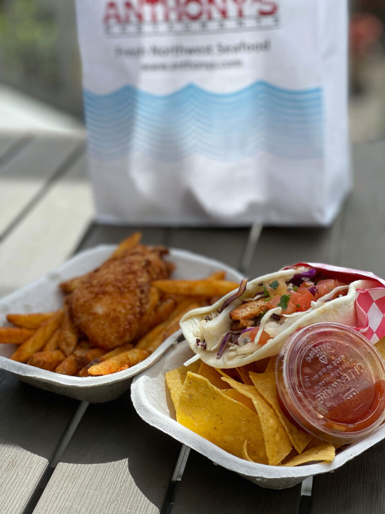 Two takeout dishes on a table: one with a fish fillet and fries, and the other with a taco, tortilla chips, and salsa. A branded paper bag in the background.