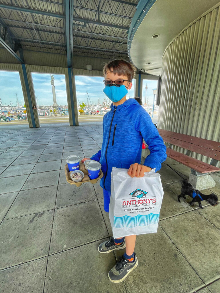 A person in a blue outfit and face mask stands under a pavilion, holding a bag labeled "Anthony's" and a tray with drinks and a dessert. A bench is on the right, and boats are visible in the background.