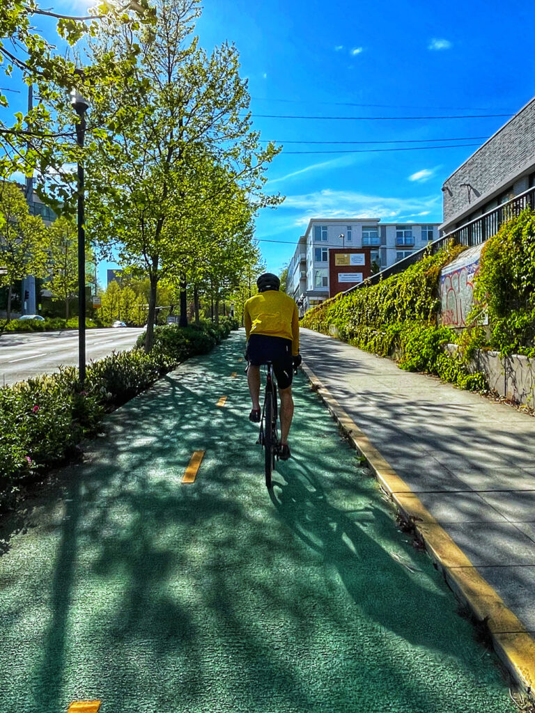 A cyclist wearing a yellow jacket rides along a green, tree-lined bike path on a sunny day. Buildings with graffiti-covered walls are on the right, while the path continues straight ahead under a clear blue sky.