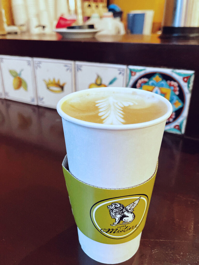 A white paper cup with a green sleeve, featuring a latte art design on top. The cup is placed on a dark wooden counter with a colorful, decorative tile backsplash depicting lemons and other patterns.