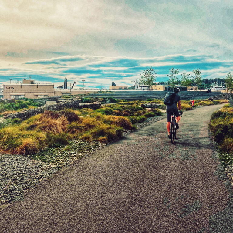 A cyclist rides on a paved path through a grassy area with industrial buildings in the background. The sky is cloudy with patches of blue.
