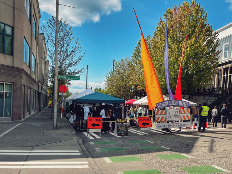 A street market scene with colorful tents and orange banners under a sunny sky. A "Street Closed" sign is visible in front of a detour sign. People browse stalls along a closed urban street lined with trees and buildings.