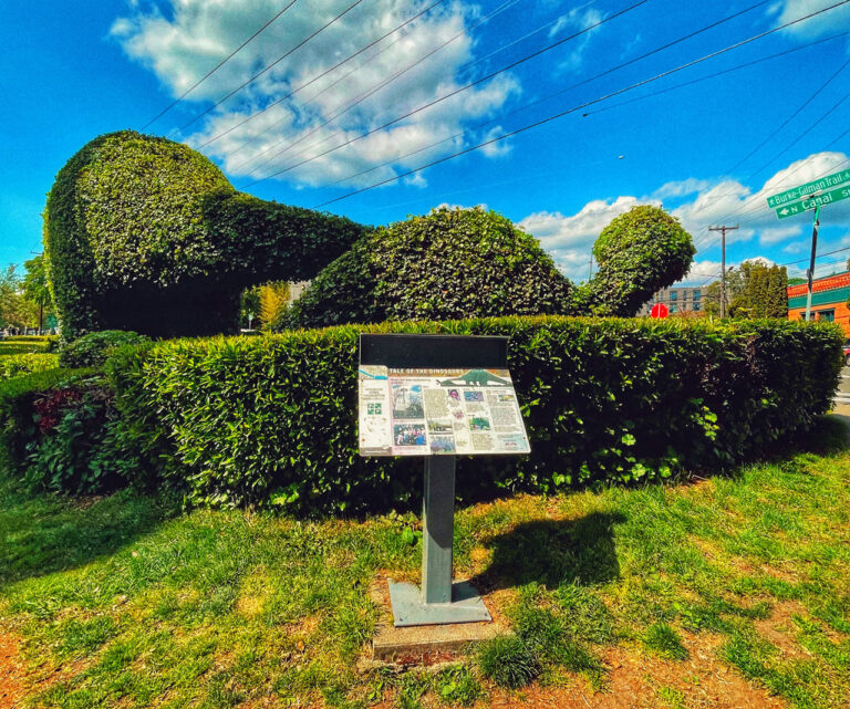 A large hedge sculpted into the shape of a dog stands on a grassy area. In front is a sign with information. Two street signs are visible on the right, and the sky is partly cloudy.