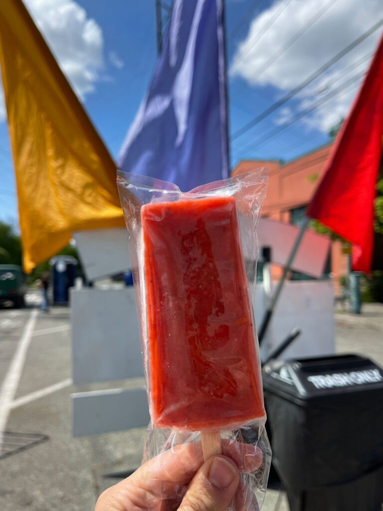 A hand holding a red popsicle wrapped in clear plastic is in the foreground. The background features colorful fabric flags—yellow, red, blue—fluttering above a sidewalk, with a blue sky and some clouds visible.