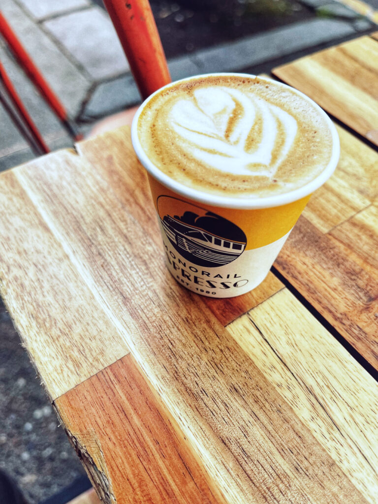 A paper cup filled with a latte featuring latte art sits on a wooden table. A part of a red chair and a glimpse of the sidewalk can be seen in the background.