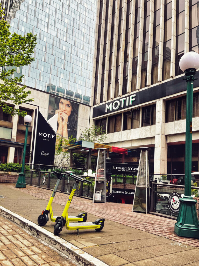 A city street scene with two yellow scooters parked on a sidewalk in front of a modern building labeled "MOTIF." A large advertisement featuring a woman is visible on the building. Trees and street lamps line the sidewalk.