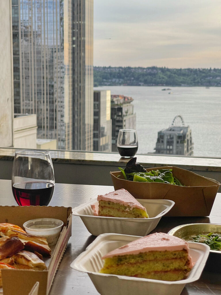 A table with plates of cake, salads, and a glass of red wine is set against a cityscape backdrop. Tall buildings, a river, and part of a Ferris wheel are visible in the distance, creating a scenic view.