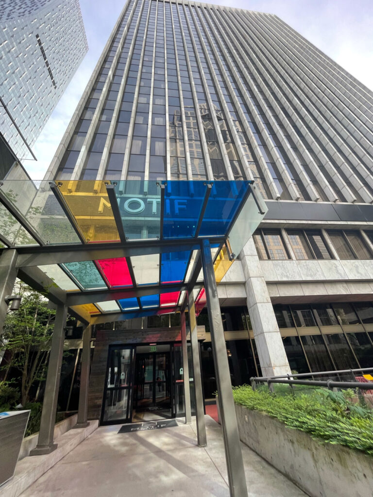 A tall, modern office building with reflective windows and a colorful glass awning in red, blue, and yellow above the entrance. Green plants line the pathway leading to the entrance under a cloudy sky.