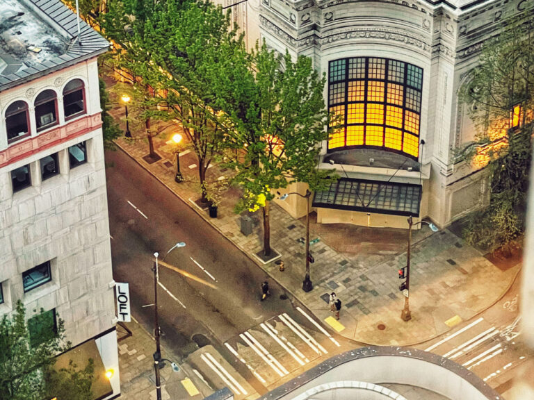 Aerial view of an urban street corner at dusk. Trees line one side of the street, and a large, illuminated window is visible on an ornate building. Streetlights cast a warm glow, and a few people walk on the pavement. The street sign reads "LOFT.