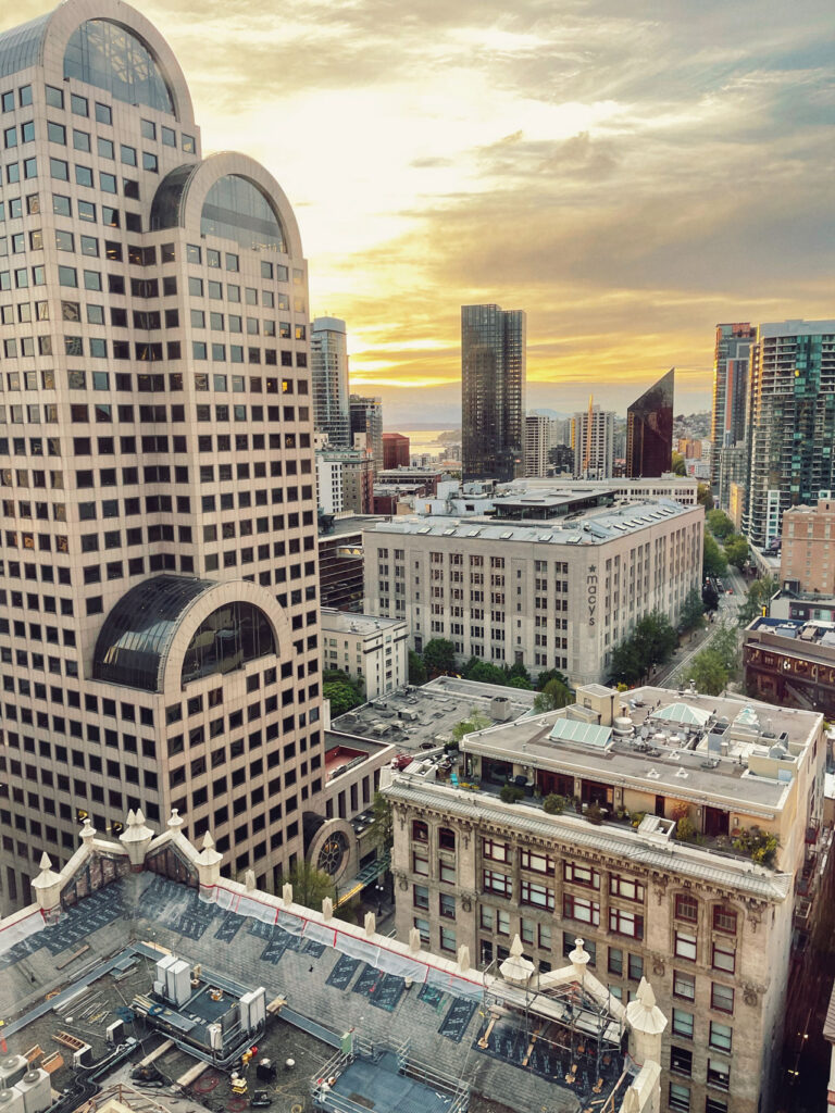 Aerial view of a cityscape at sunset, featuring modern skyscrapers and older buildings. The sky is tinged with warm hues, and the streets below are lined with trees. A large building with distinctive arched roofs stands prominently in the foreground.