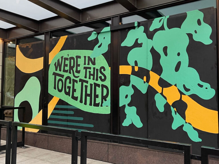 A mural with a black background features green and yellow abstract shapes. Text reads "We're in this together." The artwork is displayed on a building's exterior, with metal railings in front.