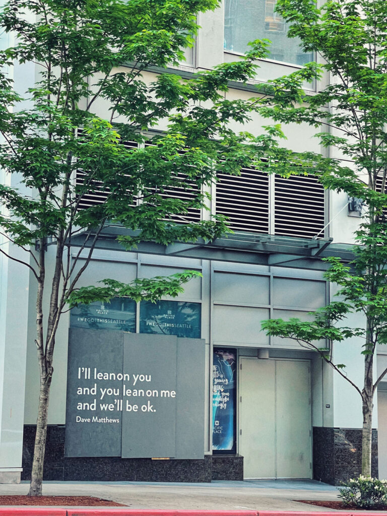 A modern building facade with a quote by Dave Matthews displayed prominently: "I’ll lean on you and you lean on me and we’ll be ok." The entrance is surrounded by green trees and the sidewalk is visible.