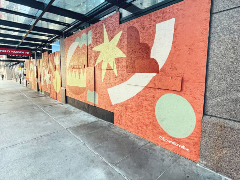Sidewalk view of a building facade with colorful abstract mural art featuring geometric shapes in orange, red, white, and green tones. The mural includes stars, circles, and angular forms, signed by "@ocreative" at the bottom right.