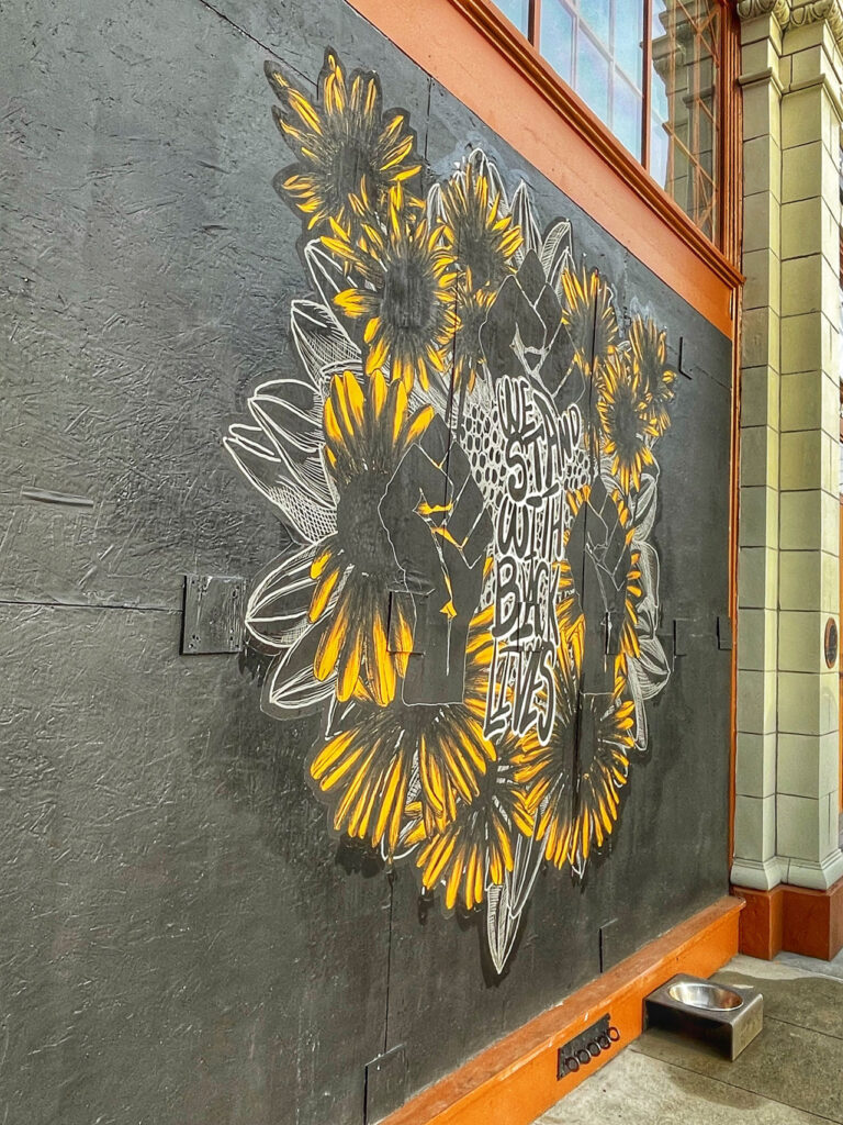 Mural featuring stylized sunflowers painted on a dark wall. Text in the middle reads "WE SHOULD OUTTA BEGI LYFS." The art is bordered by an orange frame and a metal dog bowl is on the ground below.