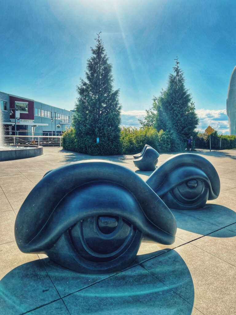 Sculptural installation of large, black eye shapes on a sunny plaza with modern buildings and tall green trees in the background. Shadows are cast on the pavement, and the sky is bright with scattered clouds.
