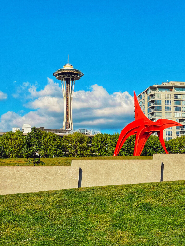 A bright, sunny day in Seattle featuring the Space Needle and the "Eagle" sculpture in the foreground. The sky is clear with a few fluffy clouds, and modern buildings are partially visible behind greenery.