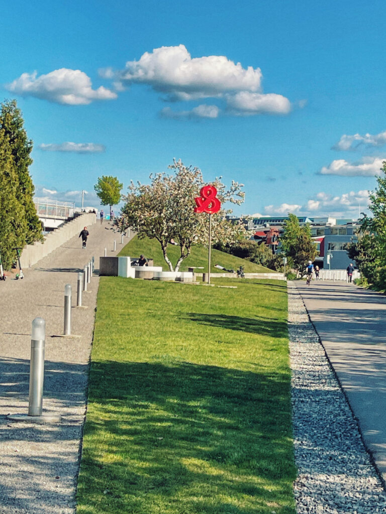 A park scene with a grassy hill and a pathway on each side. A sculpture of red roses stands near a blossoming tree under a blue sky with scattered clouds. People are walking and sitting, enjoying the sunny day.