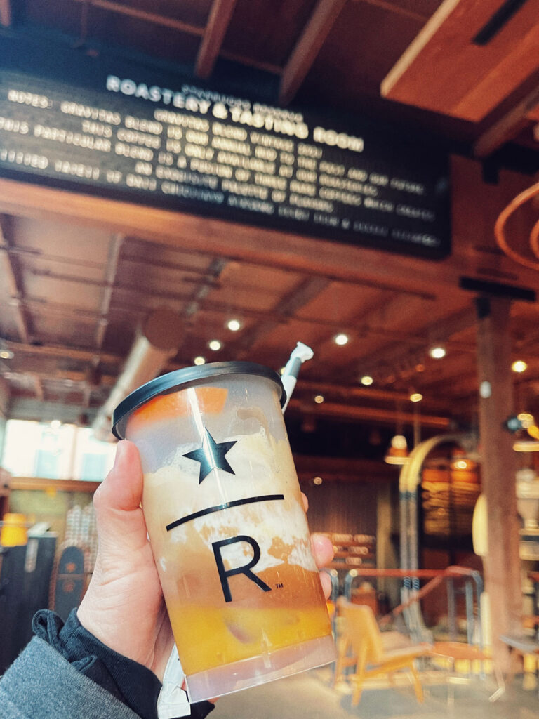 A hand holding a cold drink in a clear cup with a black star and the letter "R" printed on it. The background shows a rustic coffee shop interior with menu signage and warm lighting.