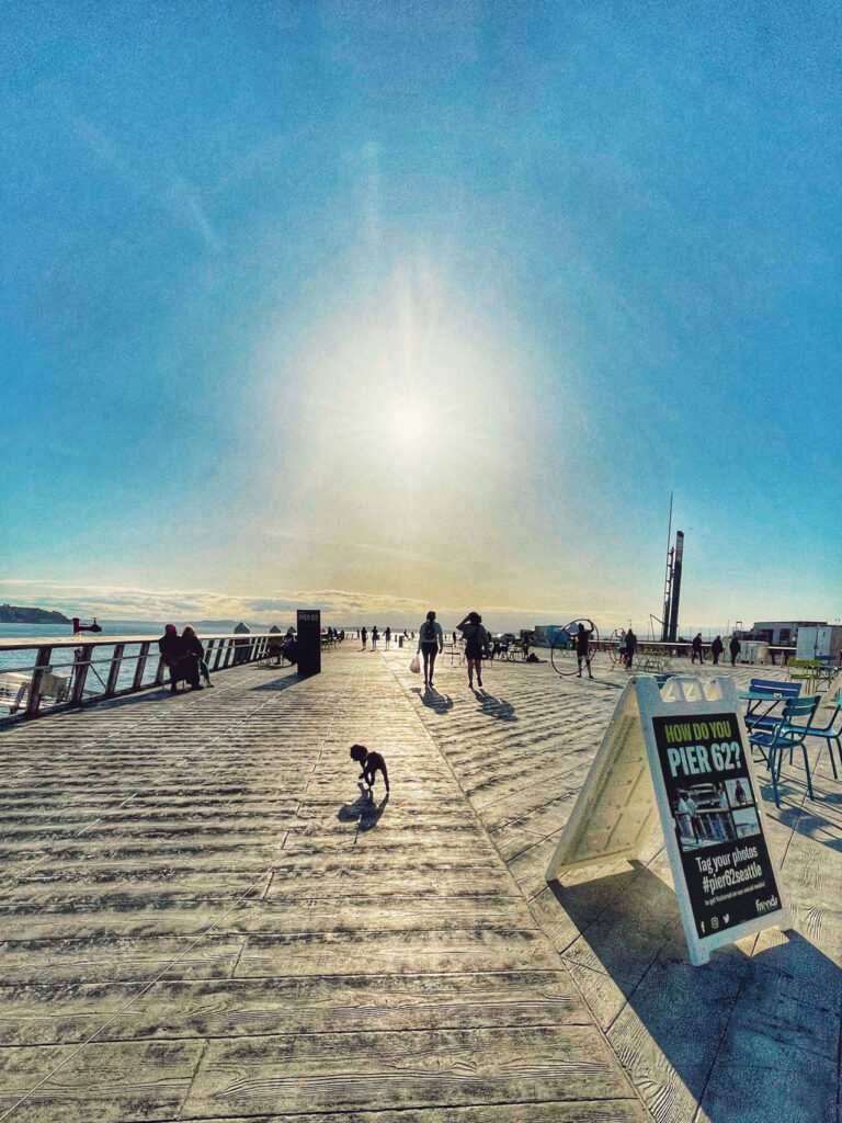 A sunny day on a wooden pier with people walking and long shadows cast by the sun. A small dog sniffs the ground, and a sign promoting photos on the right. The sky is clear and bright, creating a calm atmosphere.