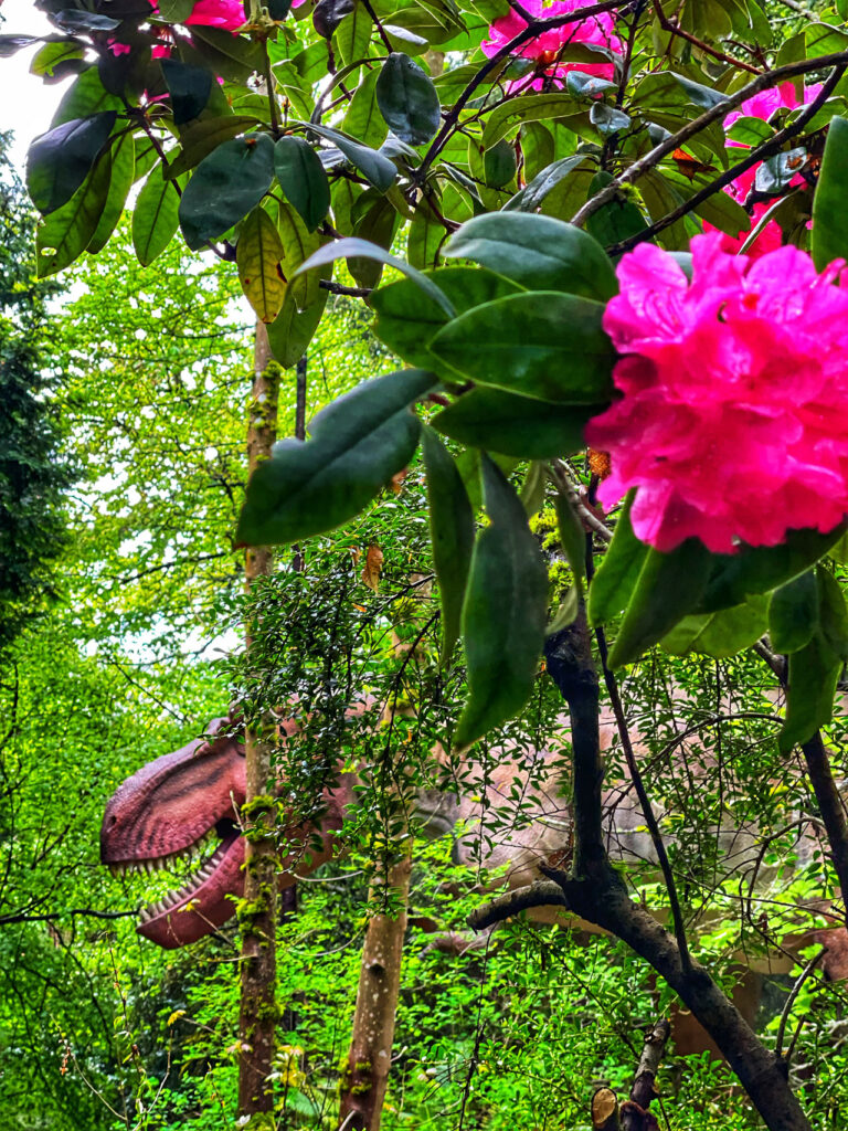 A lifelike dinosaur figure is partially hidden among lush green foliage, with vibrant pink flowers in the foreground. The creature's mouth is open, and its teeth are visible. The scene is set in a dense, forest-like environment.