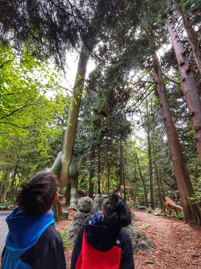 Two people in jackets look up at a large dinosaur statue in a wooded park. Tall trees surround the scene, with sunlight filtering through the branches. The dinosaur appears to be a long-necked sauropod.