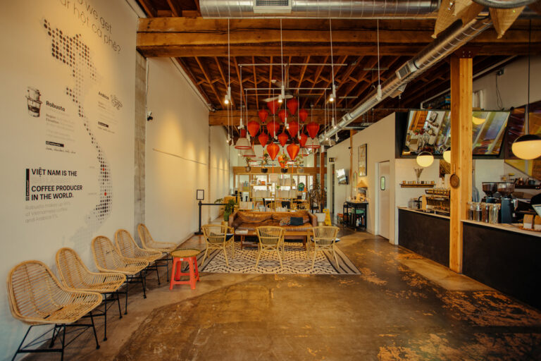 Cozy coffee shop interior with wicker chairs, a long wooden table, and red pendant lights. A mural on the left wall features coffee-related text. Exposed wooden beams and an industrial ceiling enhance the warm, inviting atmosphere.