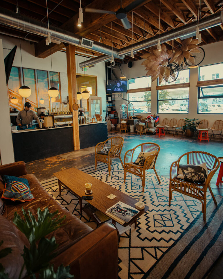A cozy coffee shop interior with wicker chairs and a leather couch around a wooden table on a geometric rug. Hanging lights illuminate the counter, where a barista is working. Large windows and rustic decor create a warm, inviting atmosphere.