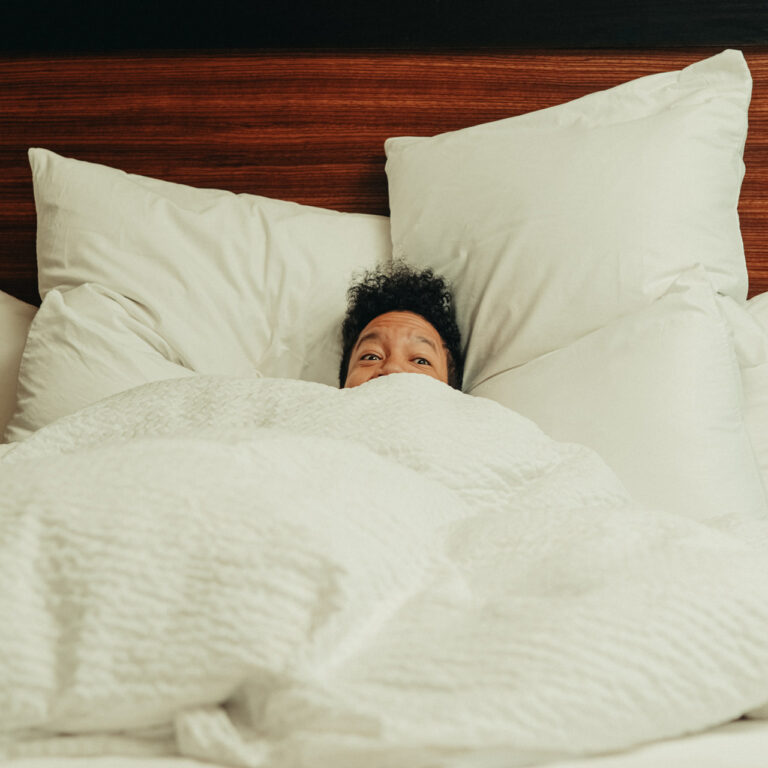 A person with curly hair is partially covered by a white comforter, only their eyes and hair visible. They are lying in bed, surrounded by several pillows, with a wooden headboard in the background.