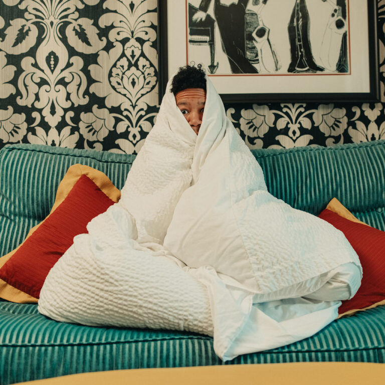 A person is wrapped in a white comforter, sitting on a teal couch with red and yellow pillows. The background features a black and white patterned wallpaper and framed artwork. They appear cozy and snug.