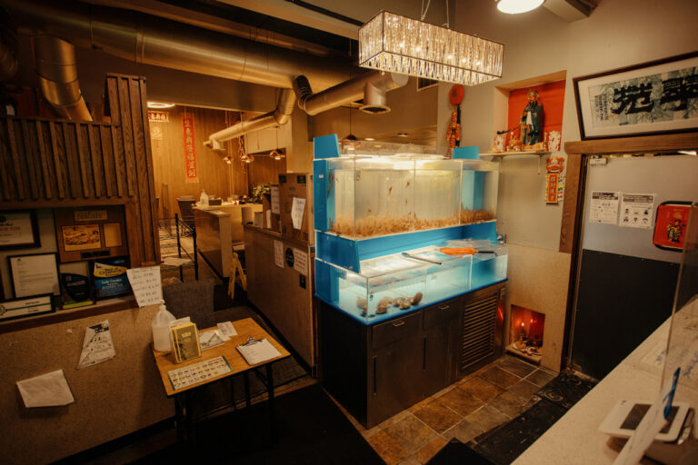 A cozy restaurant interior with dim lighting. A brightly lit blue aquarium is against the wall, containing fish and decorations. There are wooden dividers, wall art, and various papers posted on the walls and shelves. A chandelier hangs above.