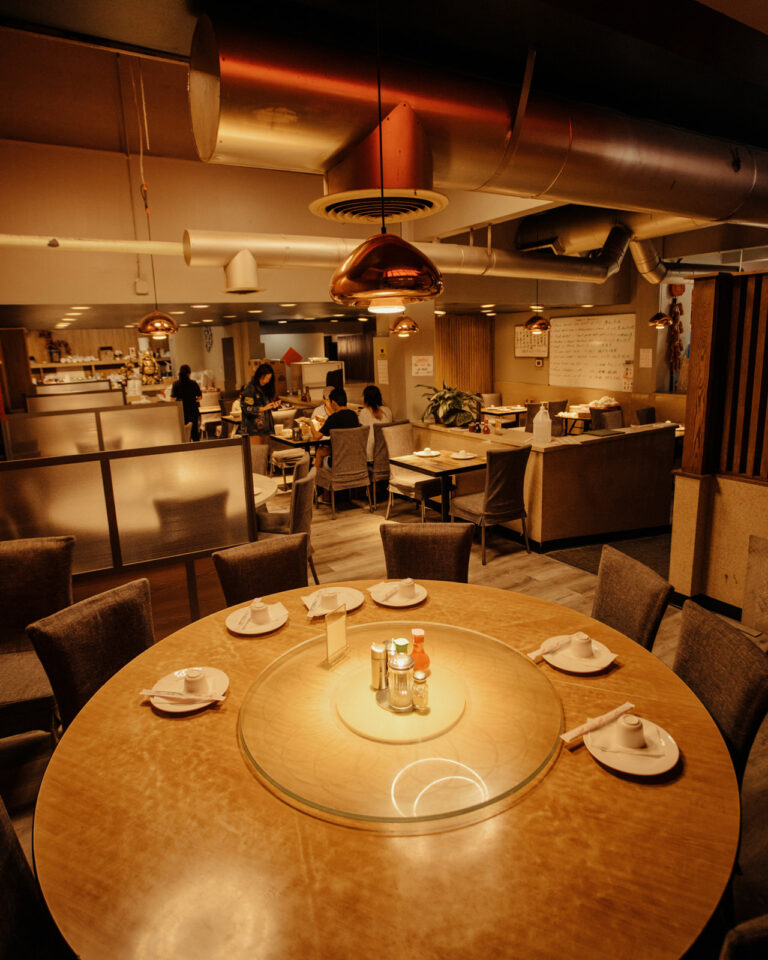 A warmly lit restaurant interior with wooden tables set for dining, featuring small sauce bottles and dishware. The space is cozy with several diners visible in the background, and modern light fixtures hang from the ceiling.