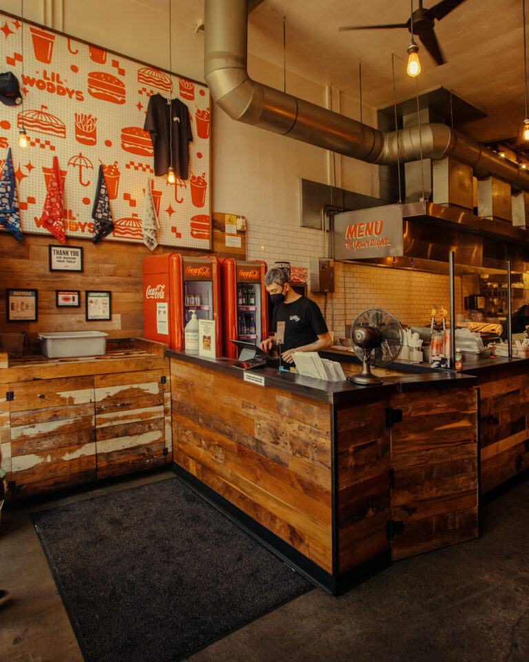 A restaurant counter with a staff member wearing a cap, surrounded by rustic wooden decor. Behind the counter are Coca-Cola dispensers and a menu sign. The wall displays various burger-themed decor and clothing items. Warm, ambient lighting fills the space.