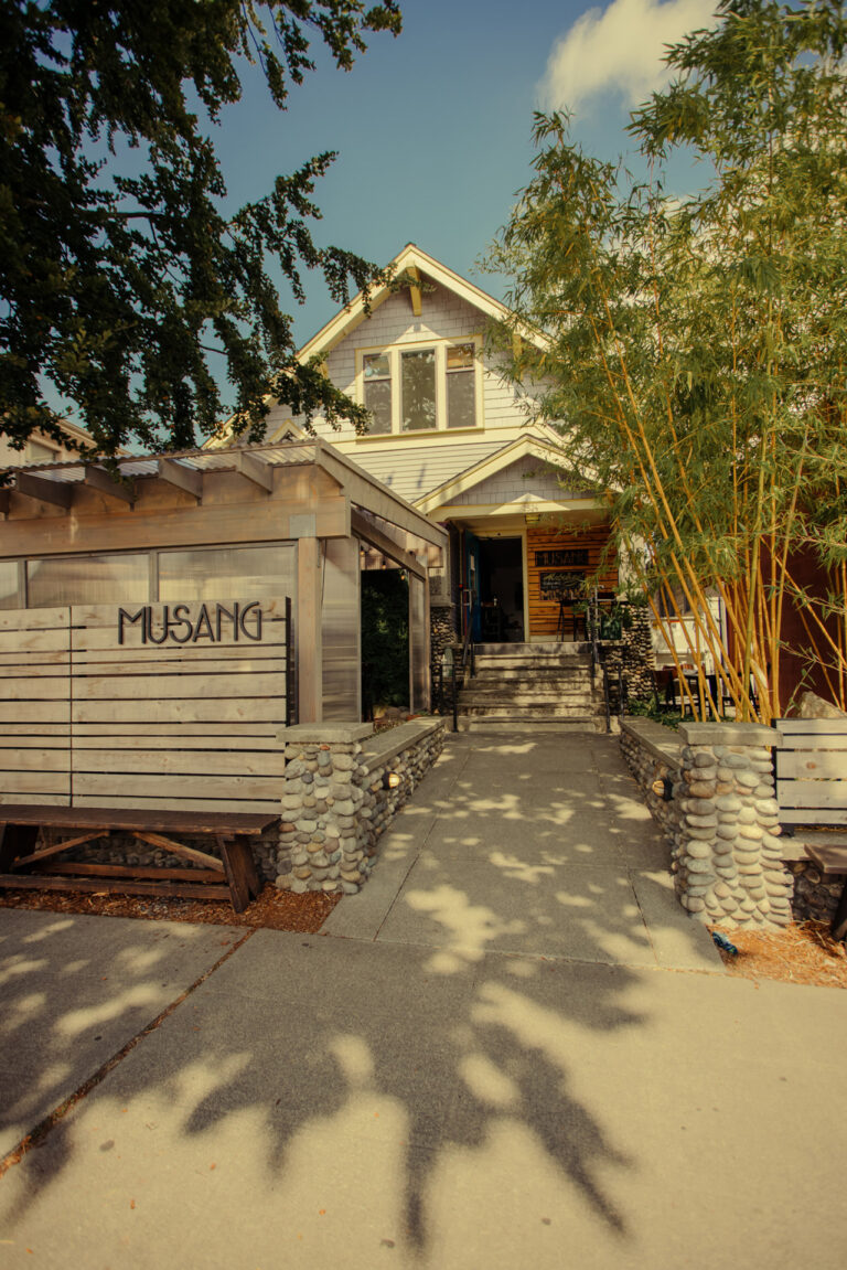 Exterior of a restaurant named Musang, featuring a rustic wooden facade and stone pathway, with bamboo plants on the right. A stairway leads to the entrance of the building under a clear blue sky.