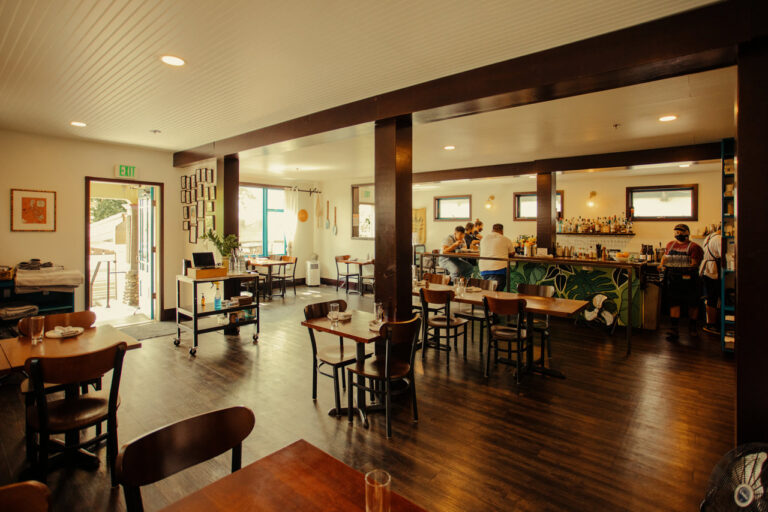 A cozy cafe interior with wooden furniture and a polished floor. The bar area is visible in the background where staff are working. Large windows let in natural light. The walls are decorated with framed art and mirrors. A door leads to an outdoor area.