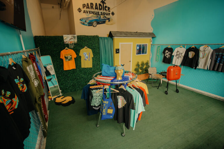 A small boutique with a tropical theme displays colorful clothing on hangers and tables. T-shirts, a grill, and decorations create a relaxed atmosphere. A sign reads "Paradise Avenue Souf," and a small model house adds charm to the setup.