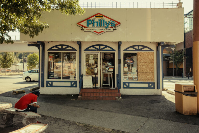 A small restaurant named "Philly's" with a sign advertising the "Best Cheesesteak in Town." The exterior is cream-colored with blue accents. A person in a red shirt sits on a nearby bench under a tree. The restaurant window displays various signs.