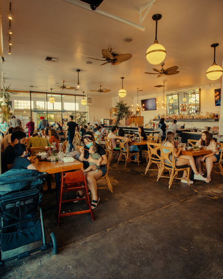 A busy café scene with people seated at wooden tables. Some are enjoying meals and drinks, while others are conversing. The space is warmly lit with ceiling fans, hanging round lights, and a long bar counter on the right. A stroller is visible in the foreground.