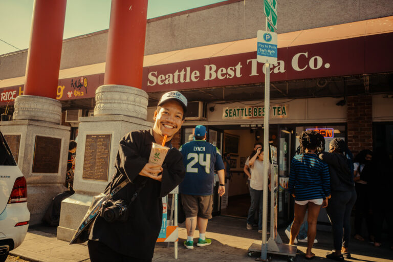 A person smiling and holding a drink in front of Seattle Best Tea Co. They are wearing a cap and a camera is hanging around their neck. A group of people is gathered outside the shop, with one person wearing a jersey with the number 24.