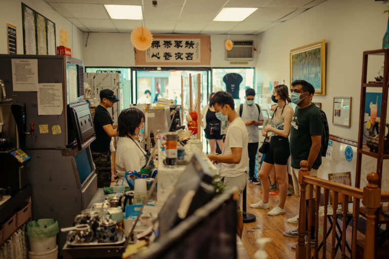 Customers line up inside a cozy café with Asian decor. Some people wear masks. A person at the counter interacts with a barista. Various items are on the counter, and a door at the back leads outside. The atmosphere is warm and bustling.