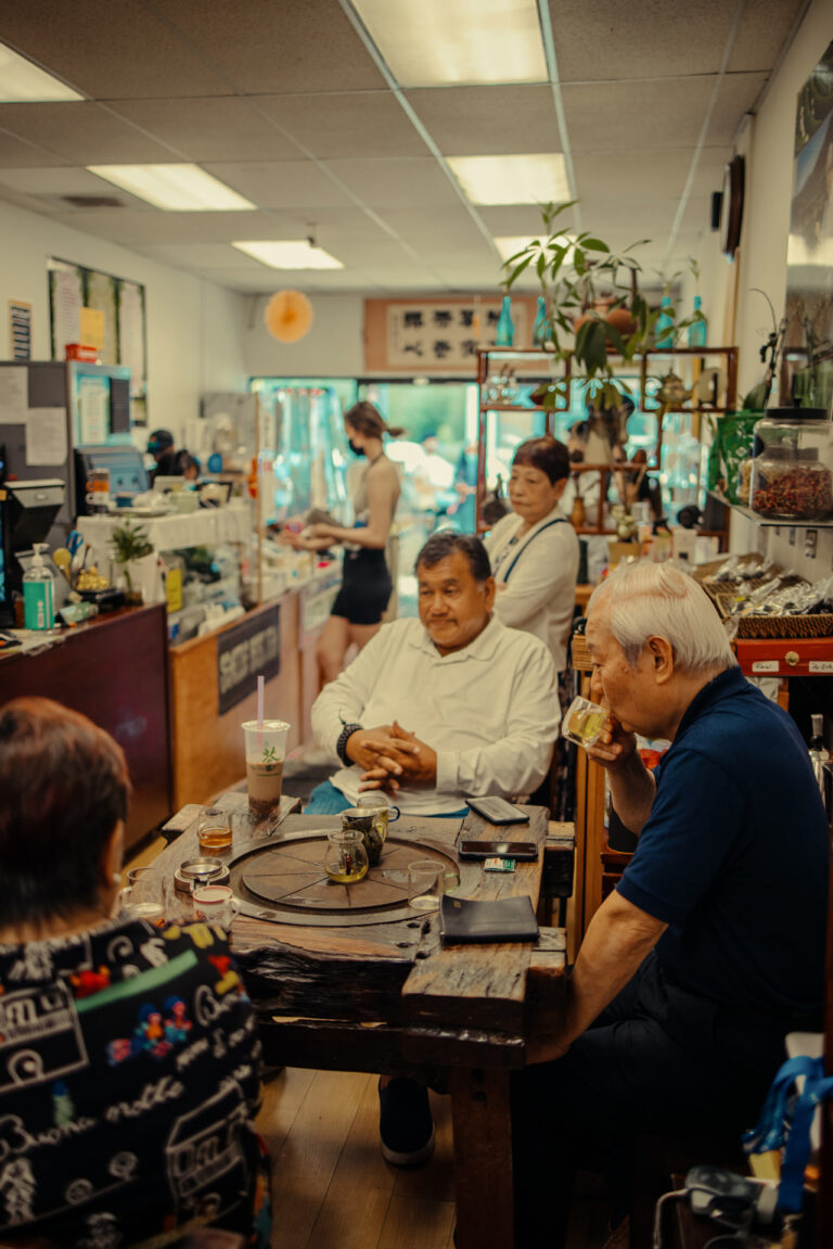 A cozy cafe with four people seated at a rustic wooden table, enjoying drinks. The interior is decorated with plants and shelves filled with various items. A barista is working in the background near the counter.