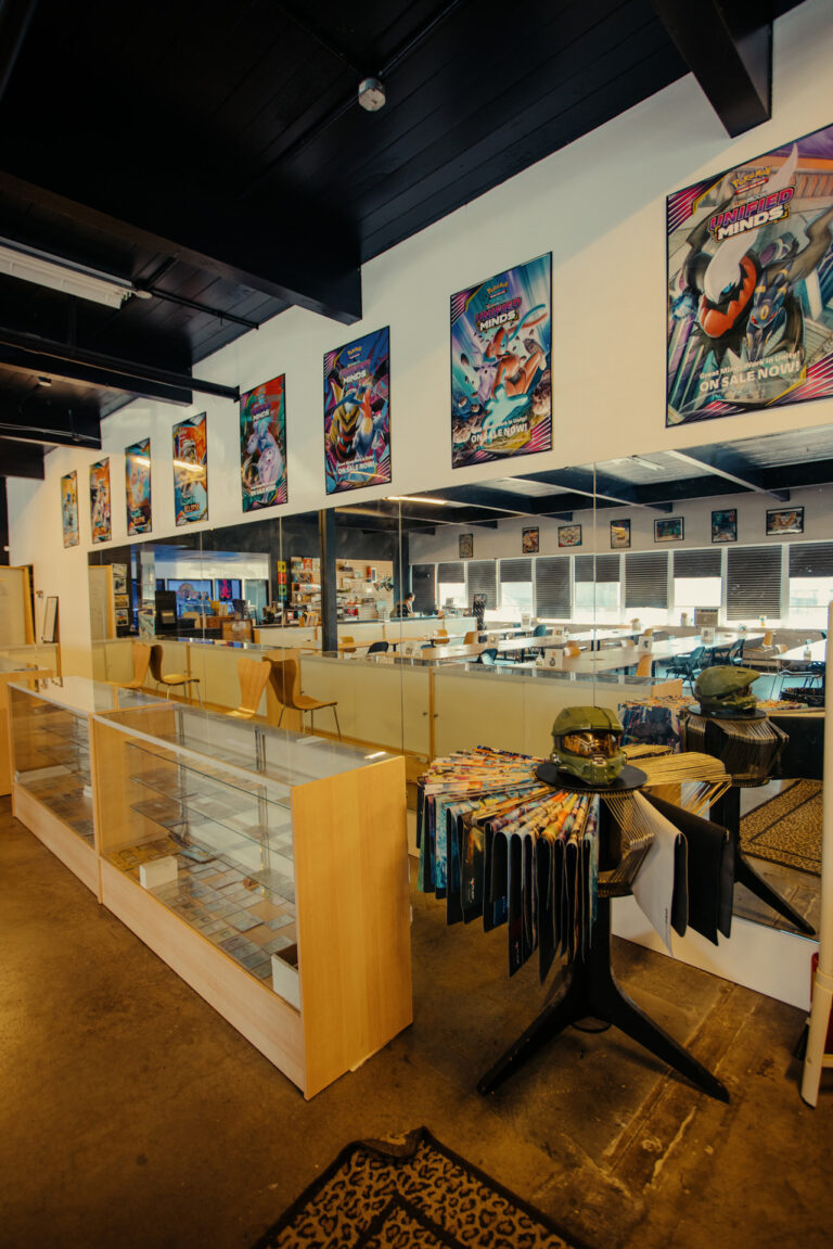 A well-lit comic book store interior featuring a long wooden display case, posters on the walls, and a circular rack filled with comic books. The room has a polished concrete floor and a counter with chairs in the background.